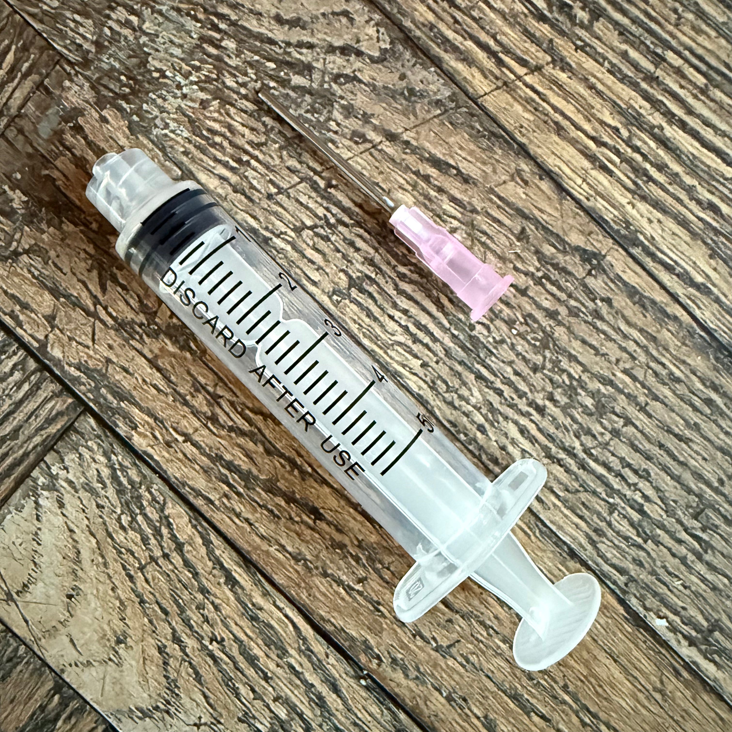5 mL ink syringe beside its detached pink-hub blunt-tip needle on a wood surface