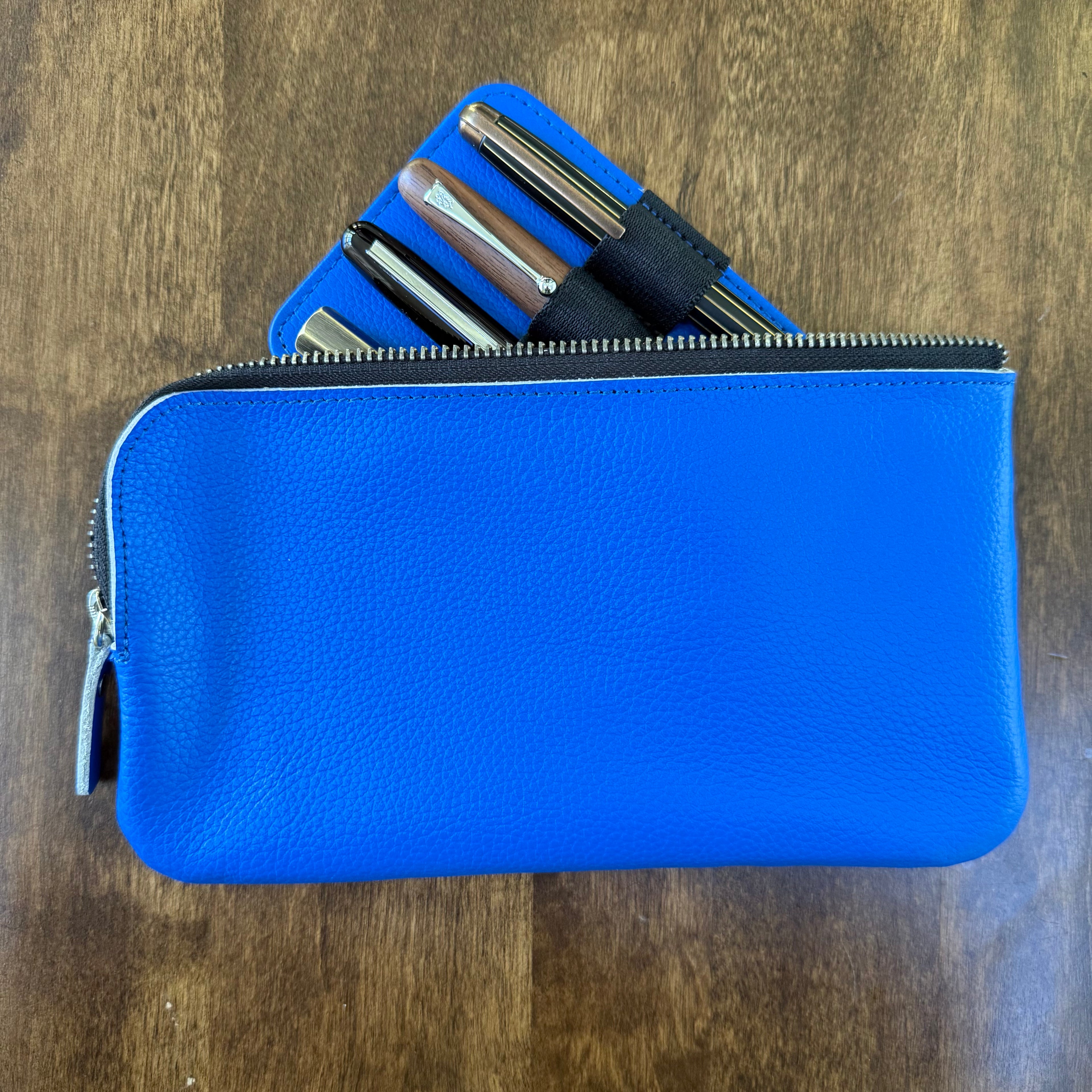 Blue cowhide leather pen case with a four-pen insert partially lifted from the open zipper pouch.