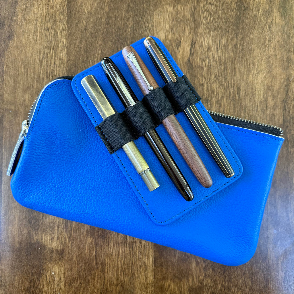 Four pens held by black elastic loops on a removable blue leather insert resting diagonally across the pouch.