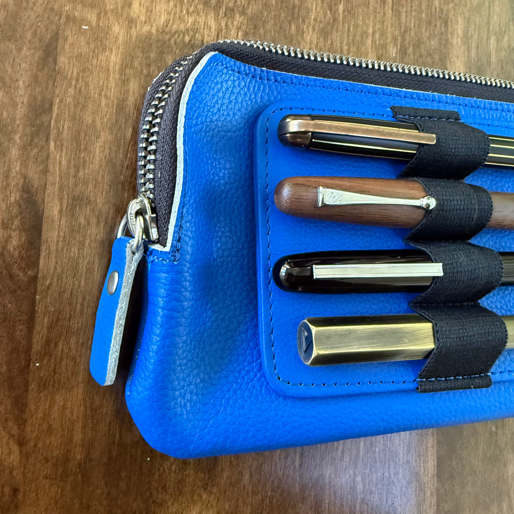 Close-up of the blue leather grain, stitching, zipper pull, and elastic loops holding four pens.
