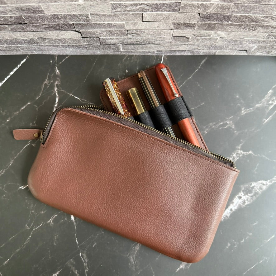 Espresso brown cowhide leather 4-pen zipper case, partly unzipped, with the pen insert and four fountain pens peeking out, on dark marble