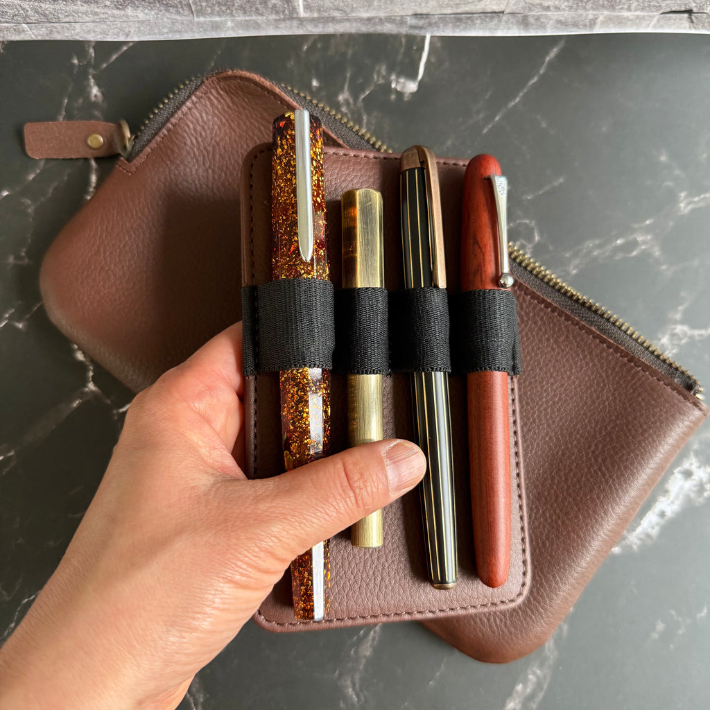 Removable leather pen insert held in hand with four fountain pens secured by black elastic loops, above the espresso brown case
