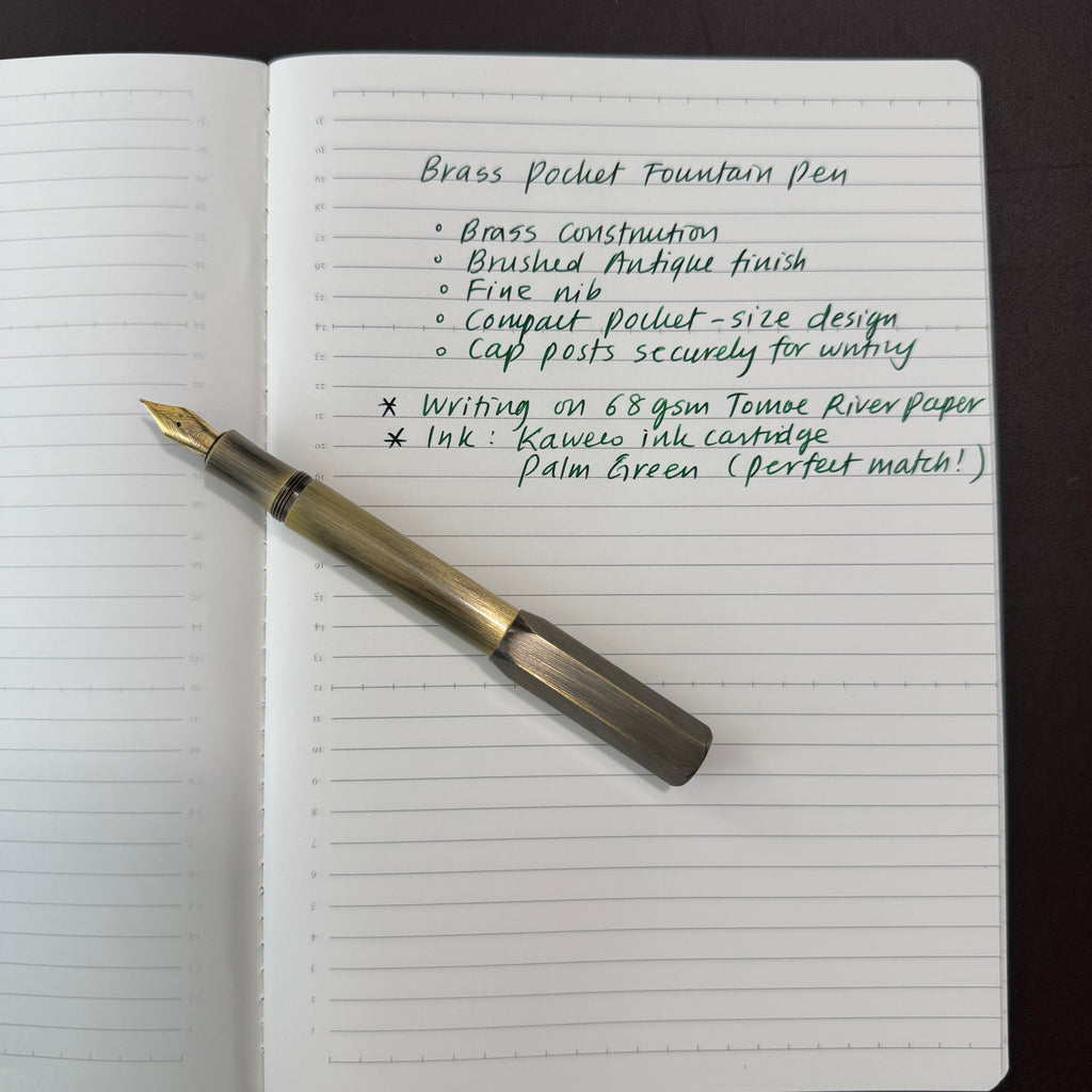 Brass pocket fountain pen with fine nib beside a handwritten writing sample in a lined notebook