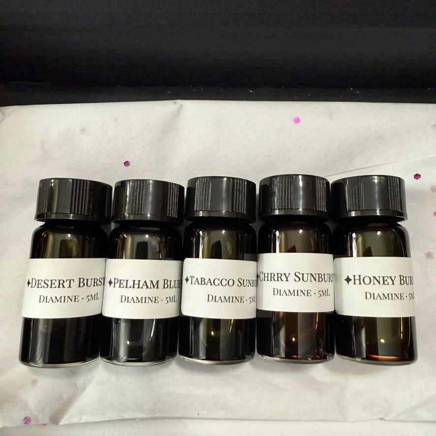 Five 5 mL glass vials of Diamine Guitar series ink with black caps and printed labels, on white tissue with purple confetti