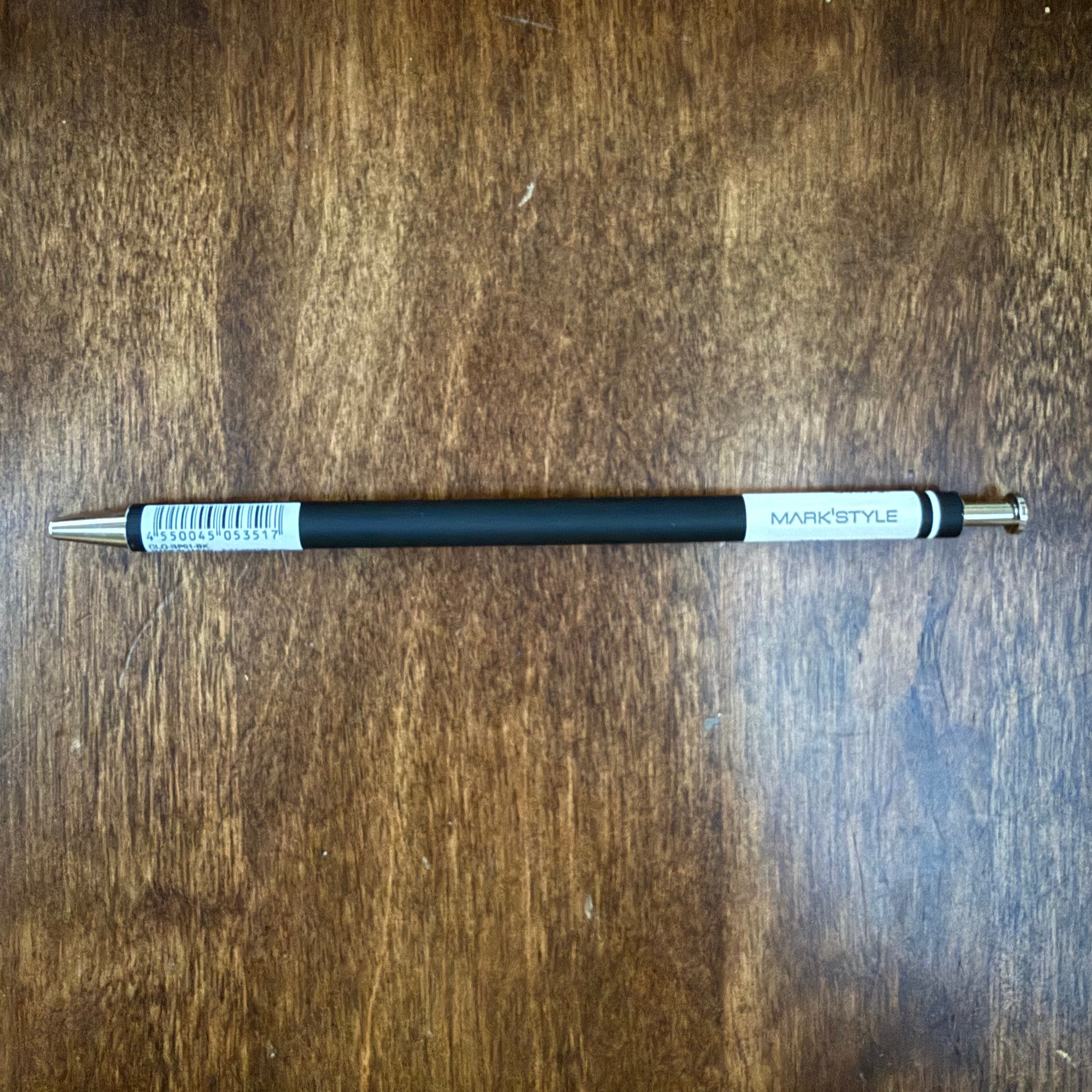 MARK’S COLORS black gel pen lying horizontally on a wooden surface, showing the white MARK’STYLE panel and silver-colored push button.