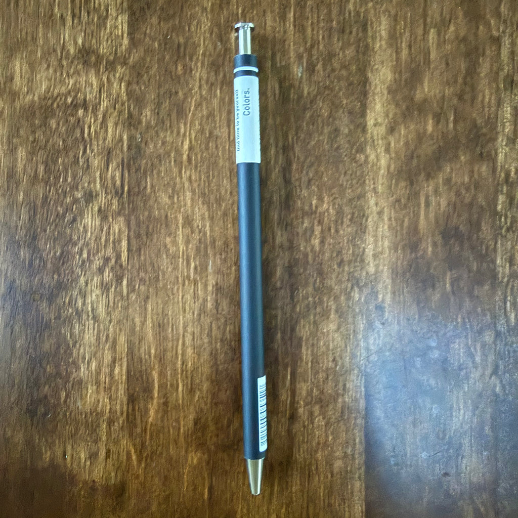 MARK’S COLORS black gel pen placed vertically on a wooden surface, showing the white Colors panel, silver-colored tip, and push button.