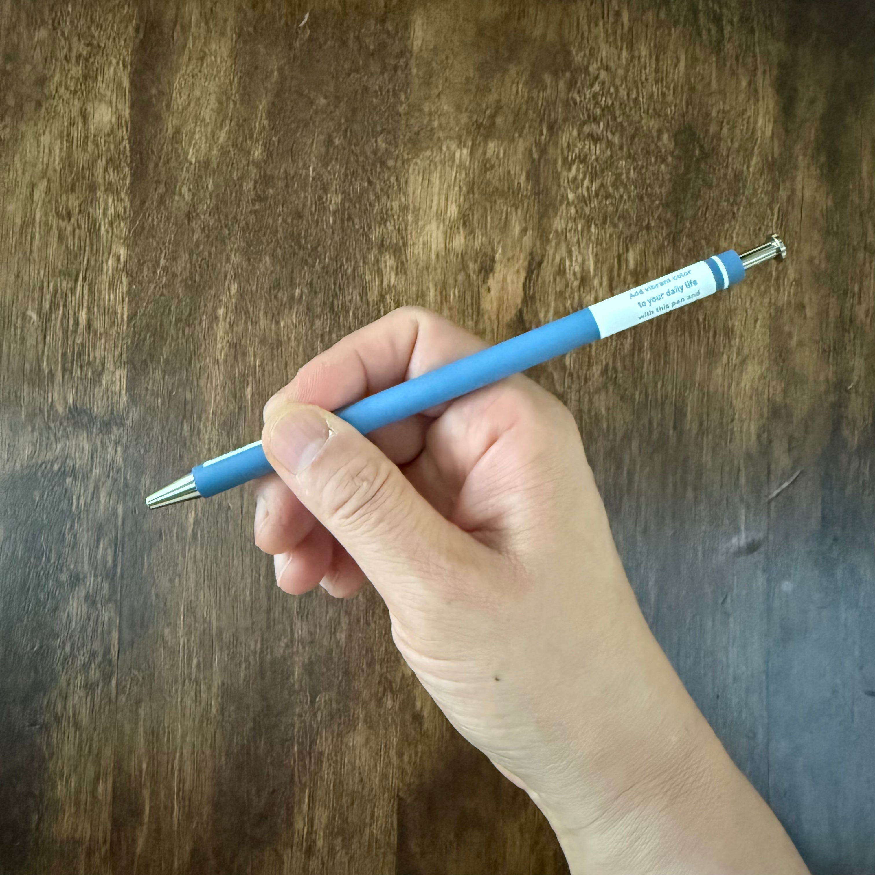 Angled view of the MARK’S COLORS blue gel pen held in a hand above a wooden surface.