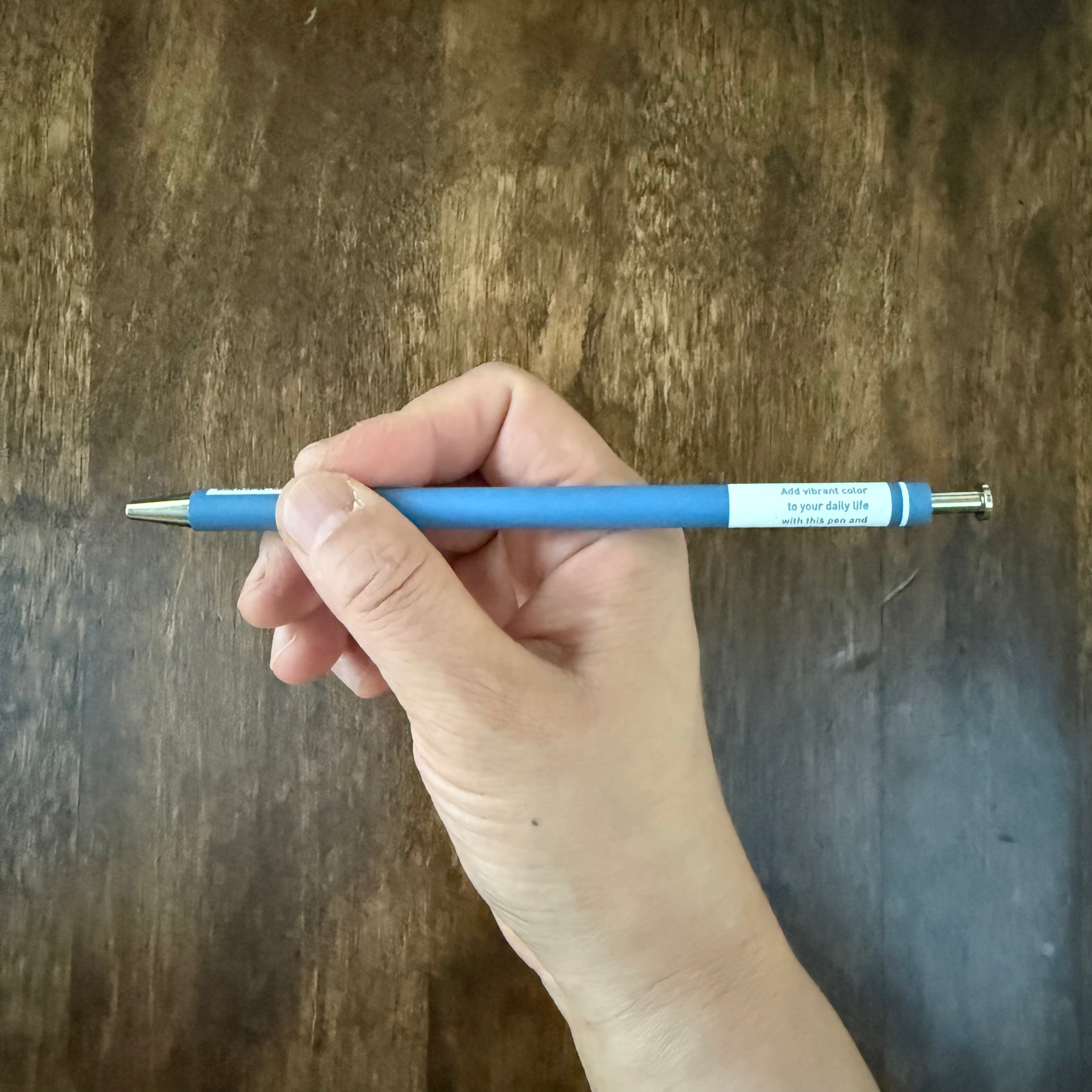MARK’S COLORS blue gel pen held horizontally in a hand, showing its slim barrel and printed white panel.