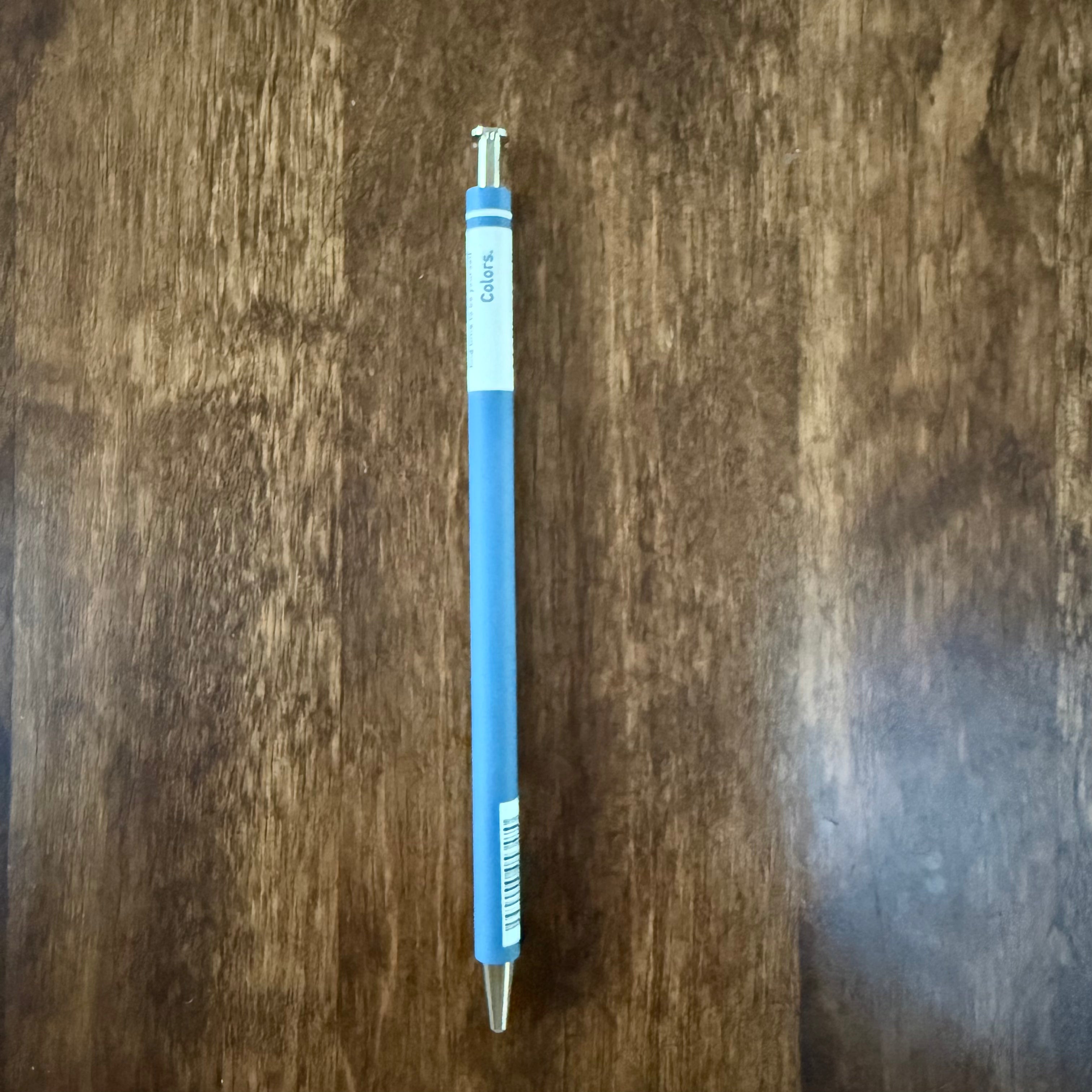 MARK’S COLORS blue gel pen placed vertically on a wooden surface, showing its white Colors panel and silver-colored tip and push button.