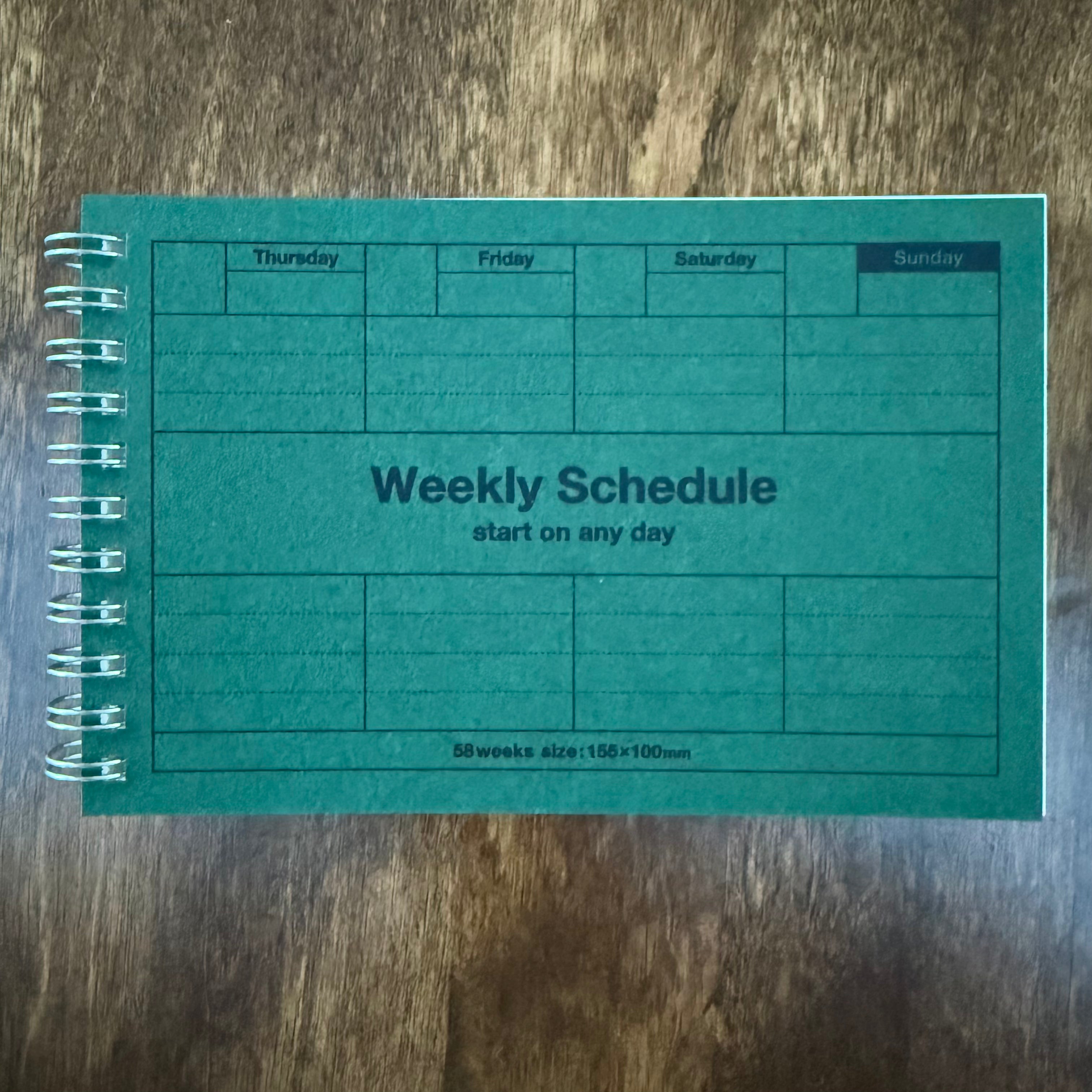 MARK’S Dayfree Dark Green weekly planner on a wooden surface, showing the Weekly Schedule cover and silver-colored wire binding.