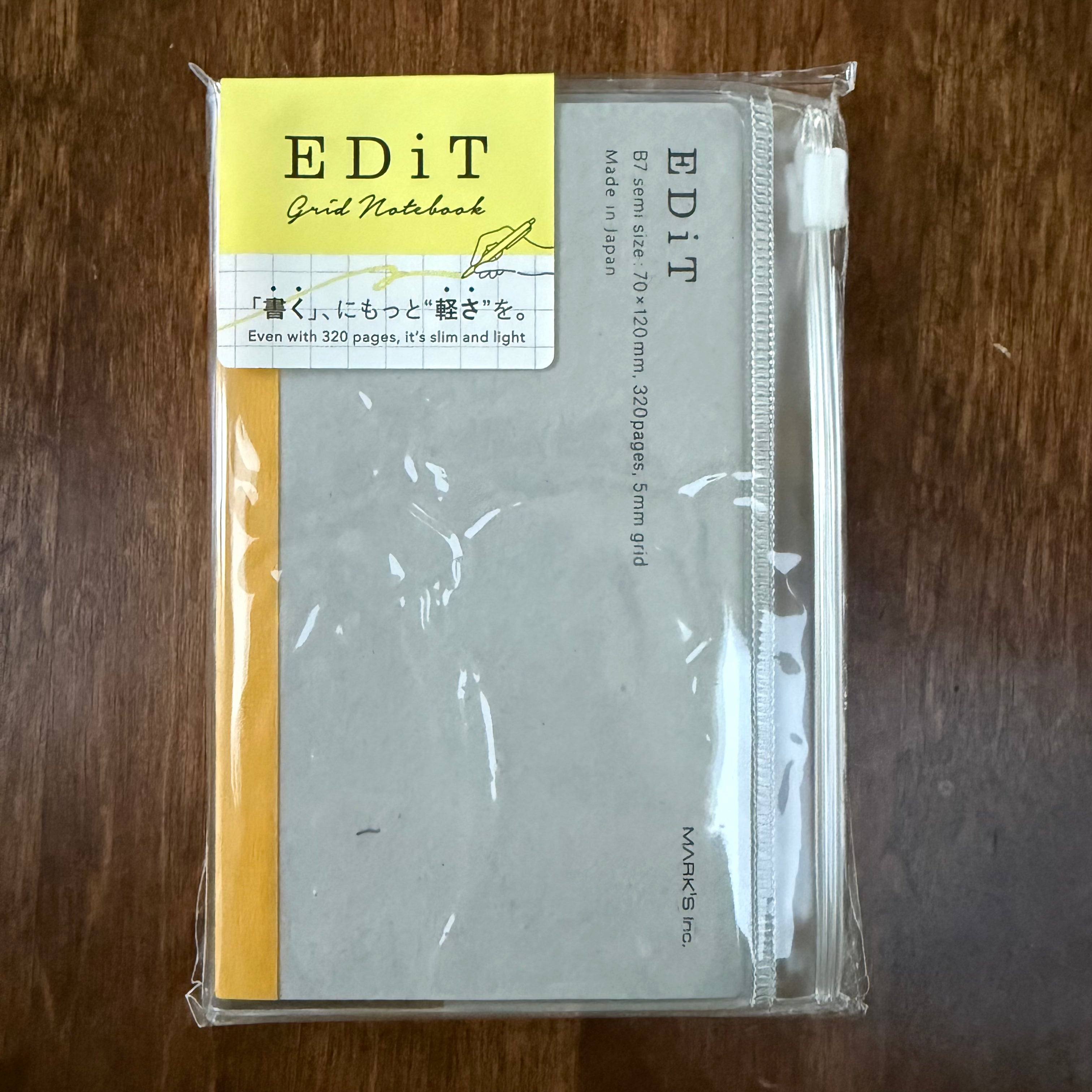 MARK’S EDiT B7 Clear notebook in packaging on a wooden surface, showing the gray cover, yellow spine, and clear zipper pocket.