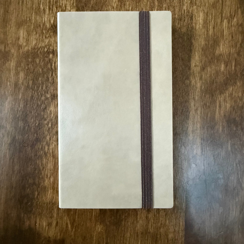 Front of the MARK’S EDiT B7 Light Latte notebook showing its beige PU cover and brown elastic closure on a wooden surface.