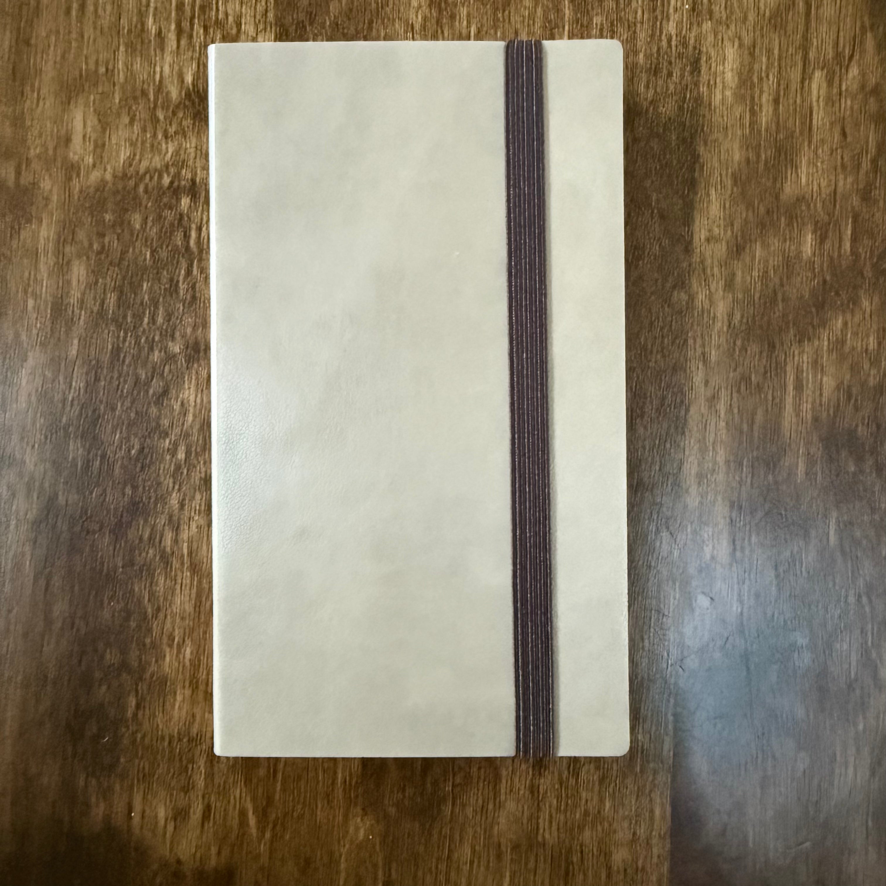 Front of the MARK’S EDiT B7 Light Latte notebook showing its beige PU cover and brown elastic closure on a wooden surface.