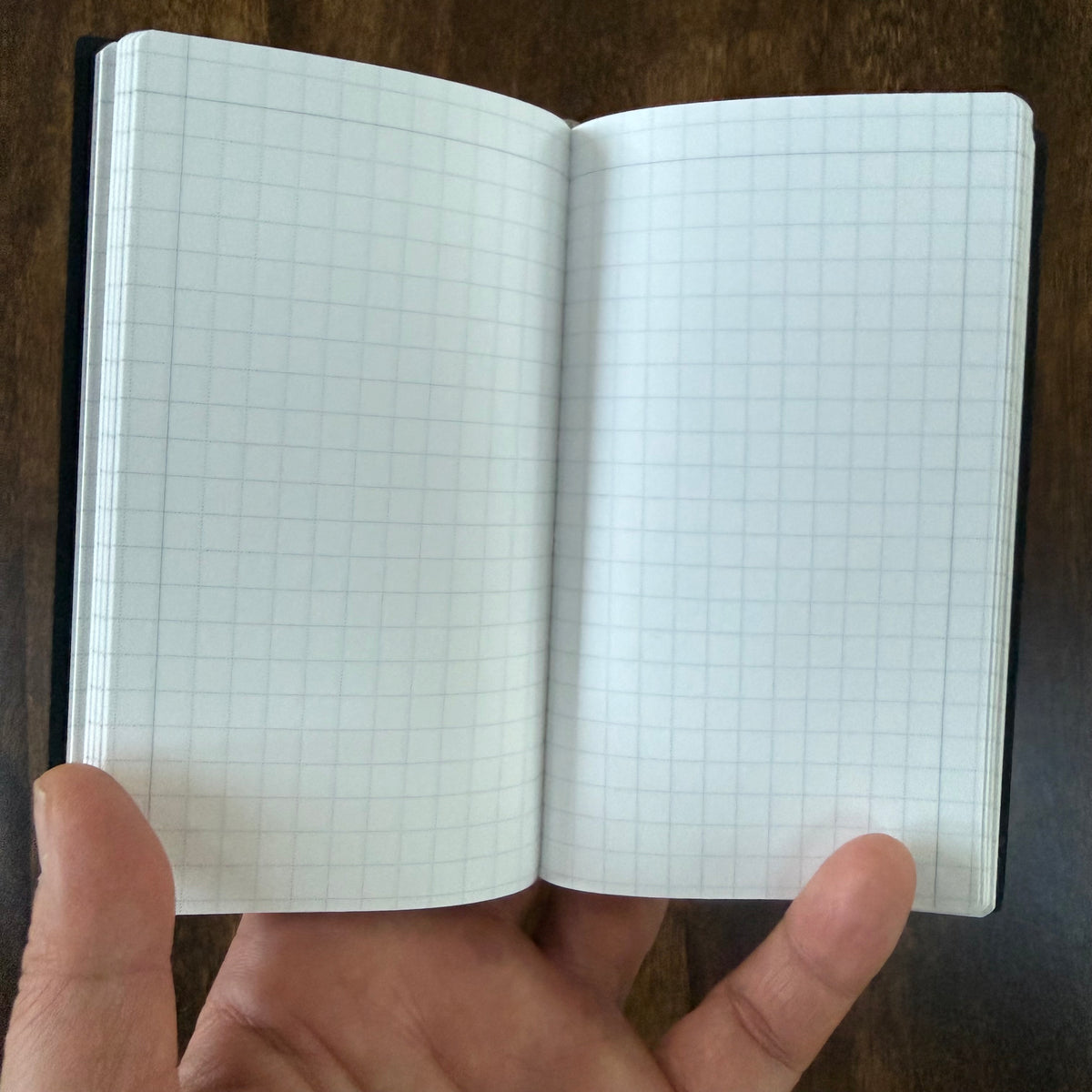MARK’S EDiT B7 notebook held open in one hand, showing its compact size and two-page spread of light gray 5 mm grid ruling.