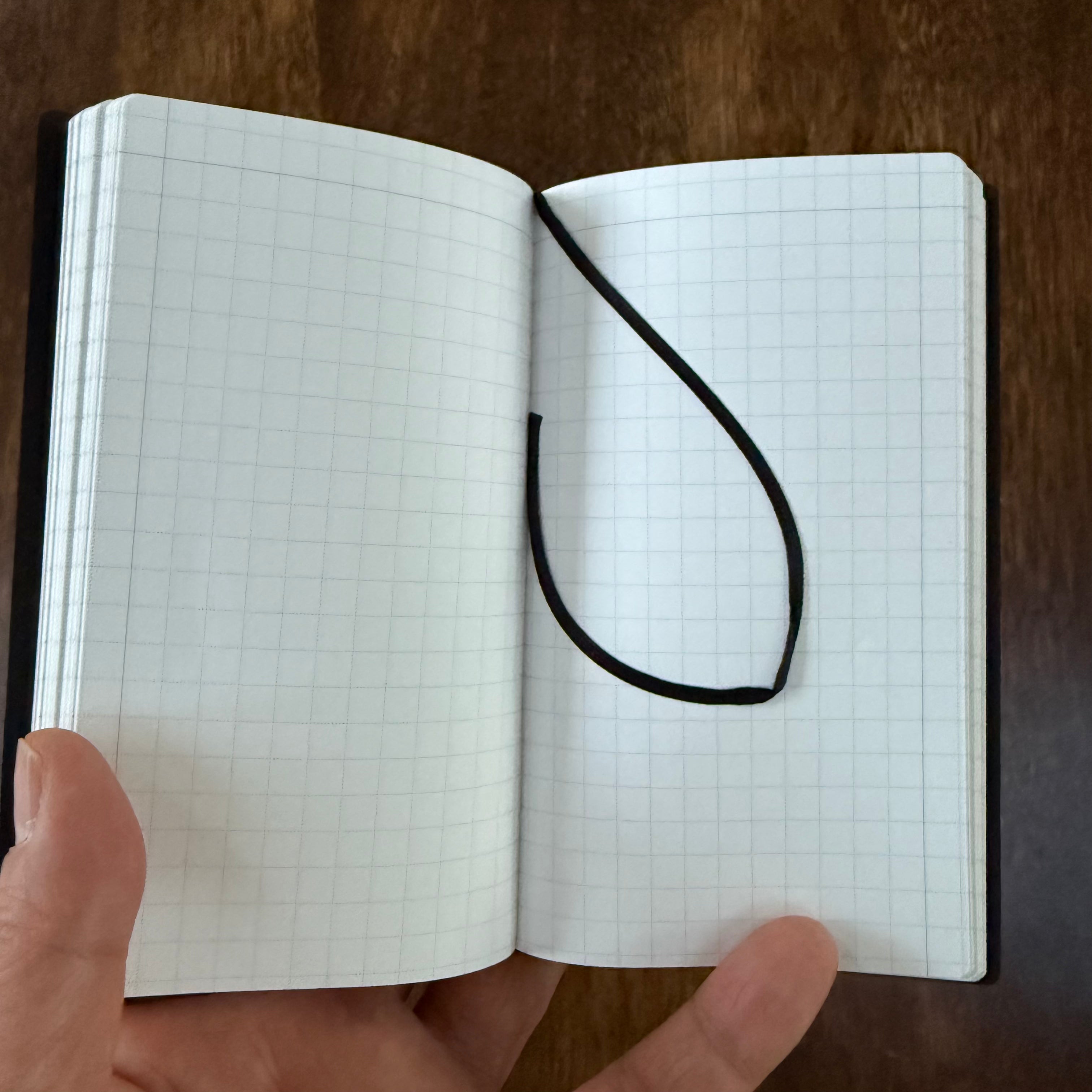 MARK’S EDiT B7 notebook held open to light gray grid pages with a dark ribbon bookmark draped across the spread.