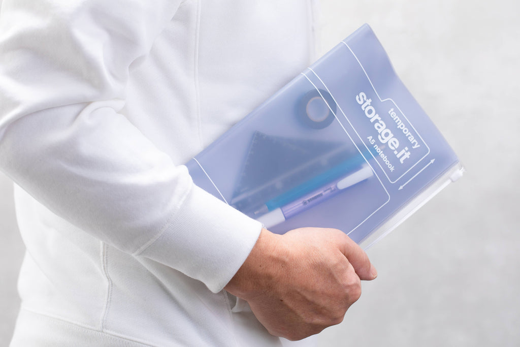 Person carrying a MARK'S Storage.it A5 notebook with pens, a ruler, and tape visible inside the translucent zip storage cover