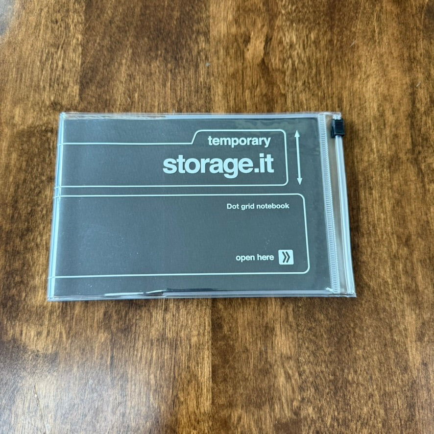 Black MARK'S Storage.it Desk Dot Grid Notebook on a wooden table, with white cover lettering and a clear slide-zip storage cover