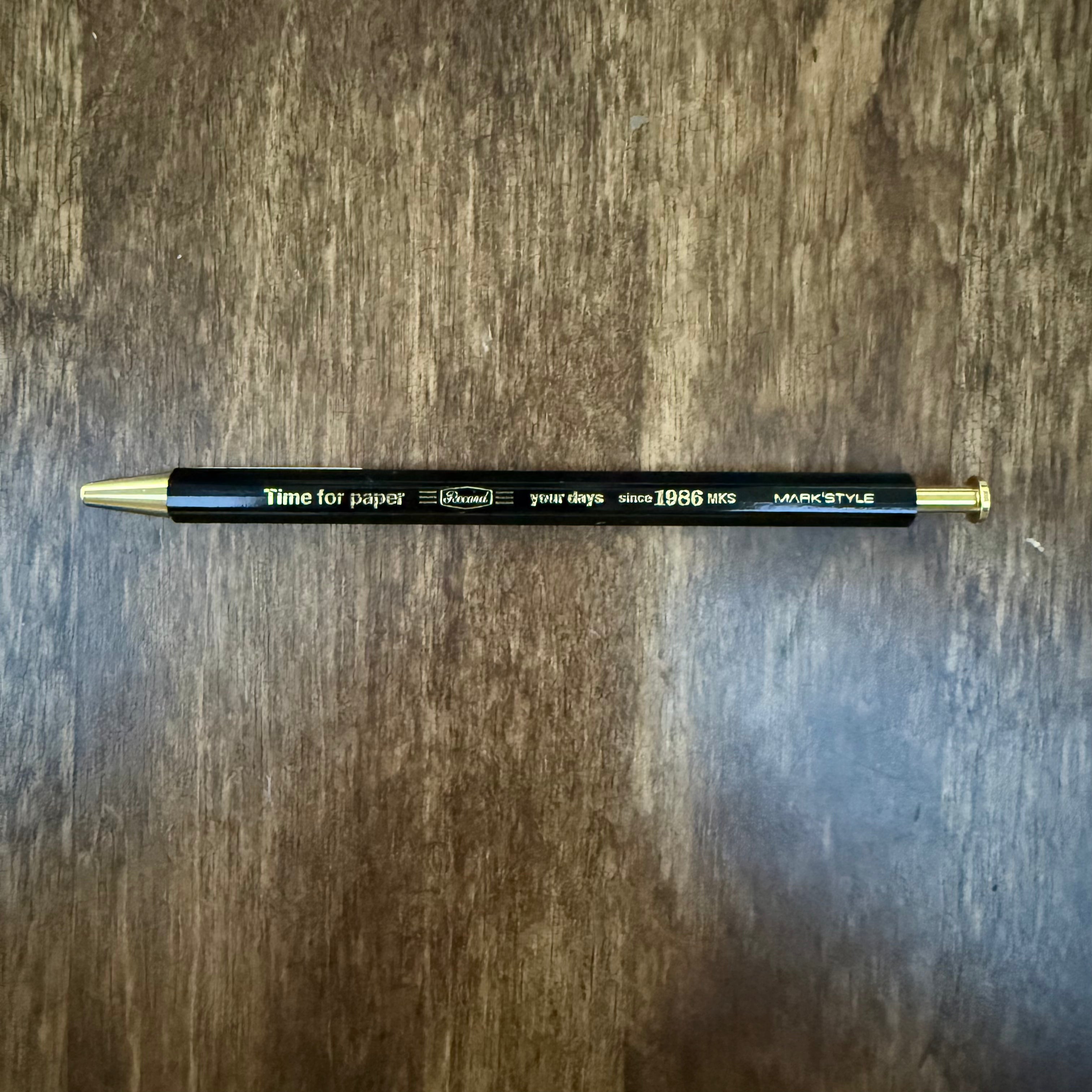 MARK’S Time for Paper black gel pen lying horizontally on a wooden surface, with gold-colored lettering, tip, and push button.