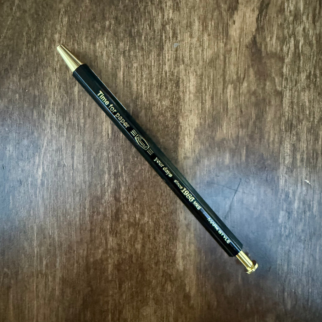 Diagonal view of the MARK’S Time for Paper gel pen, showing its slim black wooden barrel and gold-colored details.