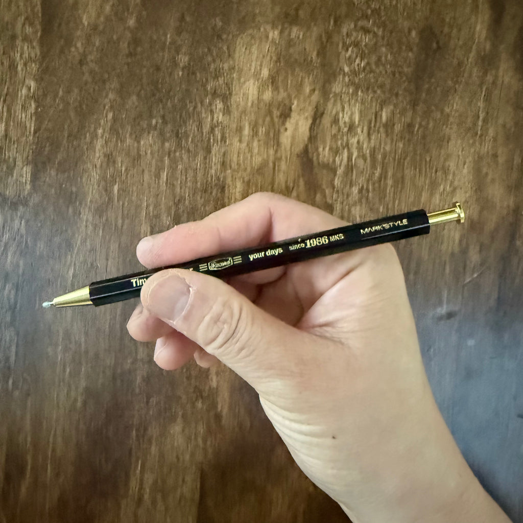 MARK’S Time for Paper black gel pen held in a hand, showing its compact size and extended writing tip.
