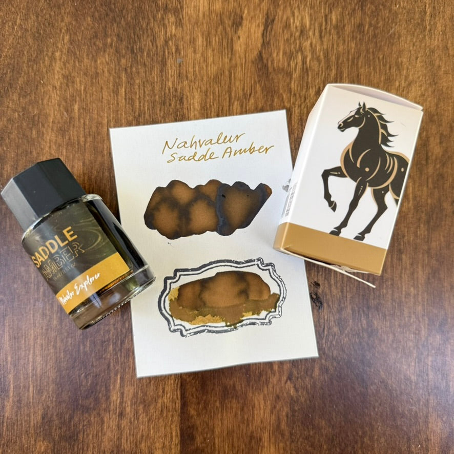 Nahvalur Explorer Saddle Amber 18 mL ink bottle beside its horse-illustrated box and two hand-painted amber-brown swatches on Tomoe River 68 gsm paper, arranged on a wooden surface.