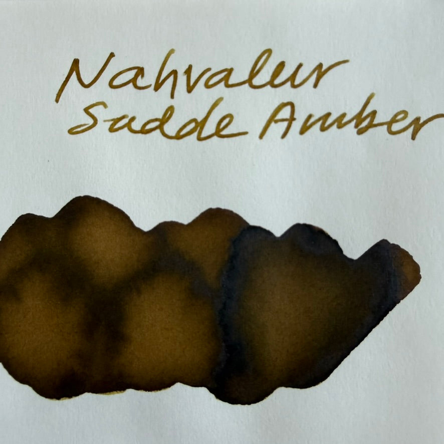 Handwritten Nahvalur Saddle Amber name above an ink swatch on Tomoe River 68 gsm paper with warm golden-brown centers and dark brown edges.