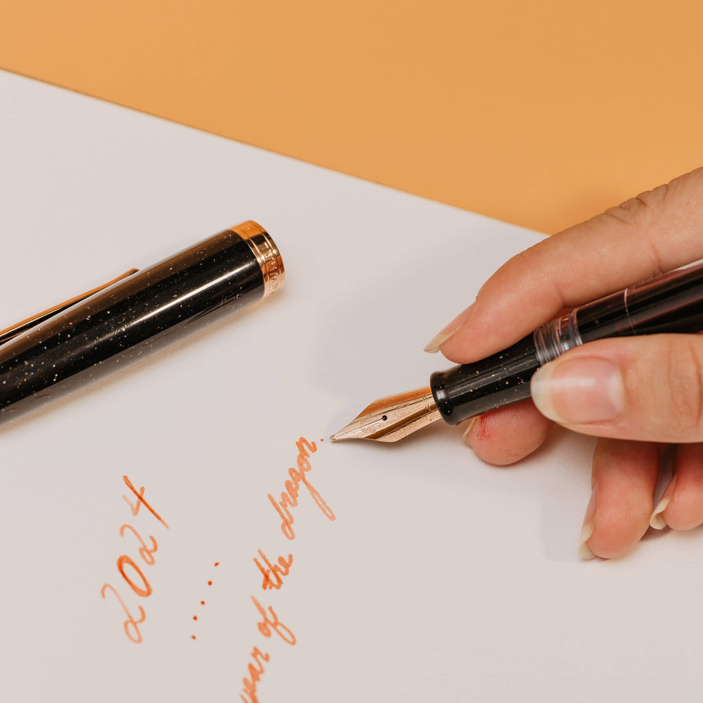 Writing in orange ink with the Nahvalur Original Plus Lovina Graphite, cap resting on the page