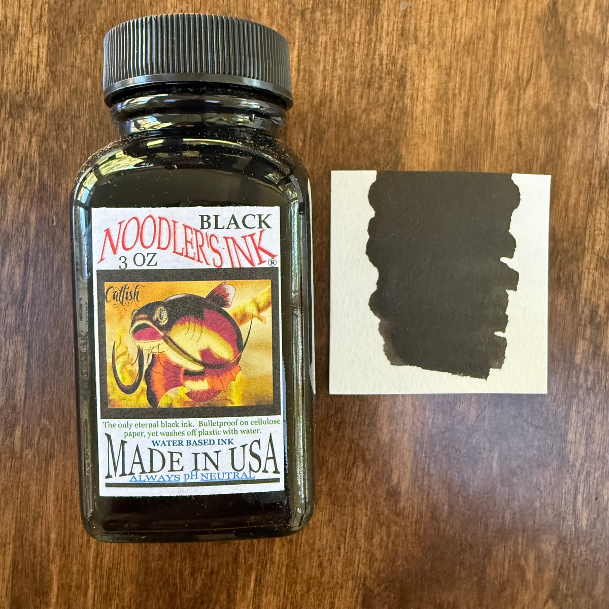 Noodler’s Black 3 oz fountain pen ink bottle with its catfish label beside a rectangular paper swatch of black ink.
