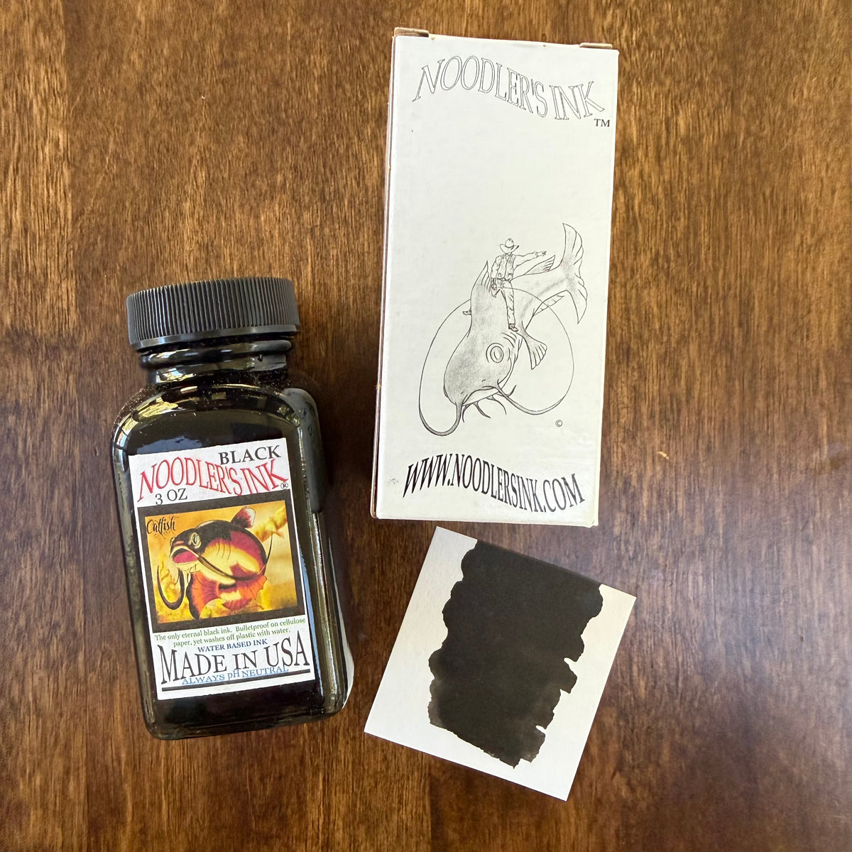 Noodler’s Black 3 oz fountain pen ink bottle beside its illustrated white box and a black ink swatch on a wooden surface.