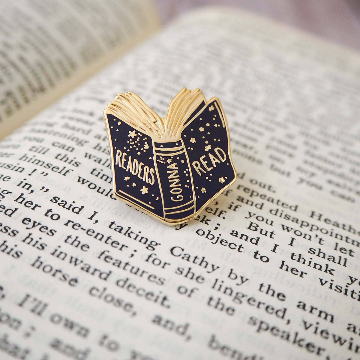 Readers Gonna Read enamel pin shaped like an open book, resting on a book page
