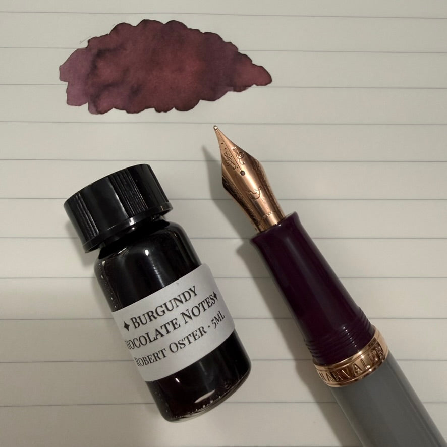 5 mL specimen bottle of Robert Oster Burgundy Chocolate Notes beside the Nahvalur Original Violet Ash fountain pen and an ink swatch