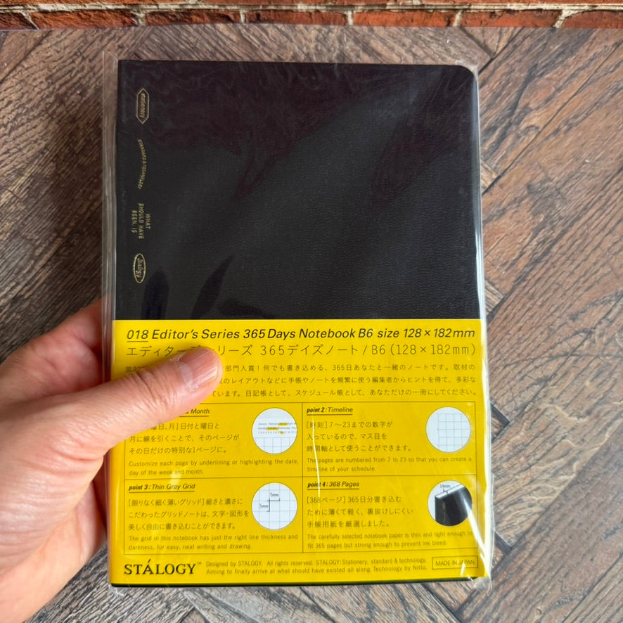 Hand holding the STALOGY 365Days B6 black notebook to show its 128 x 182 mm size
